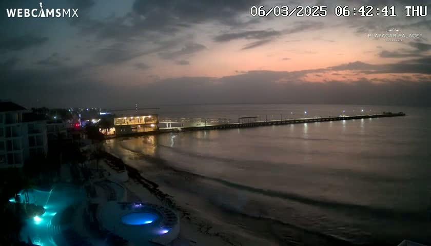 A beach at dawn, under a mostly cloudy sky with hints of pink and orange in the east, shows a long pier extending into calm water, with buildings and illuminated pools in the foreground.