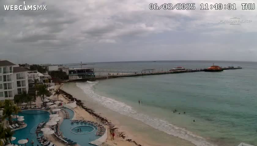 A beach with calm, light-blue water, a long pier with two boats docked, a resort with swimming pools, and an overcast sky.