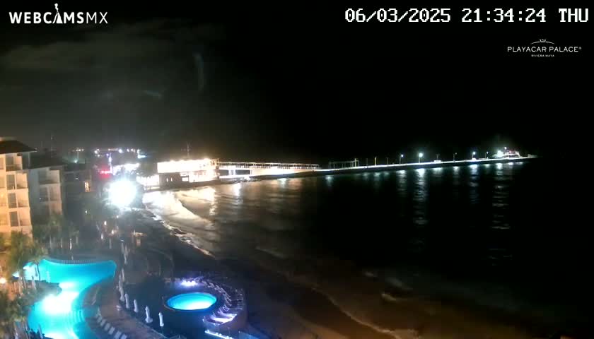 A nighttime beach scene shows a resort with illuminated pools and a pier extending into a calm, dark ocean under a mostly clear sky.