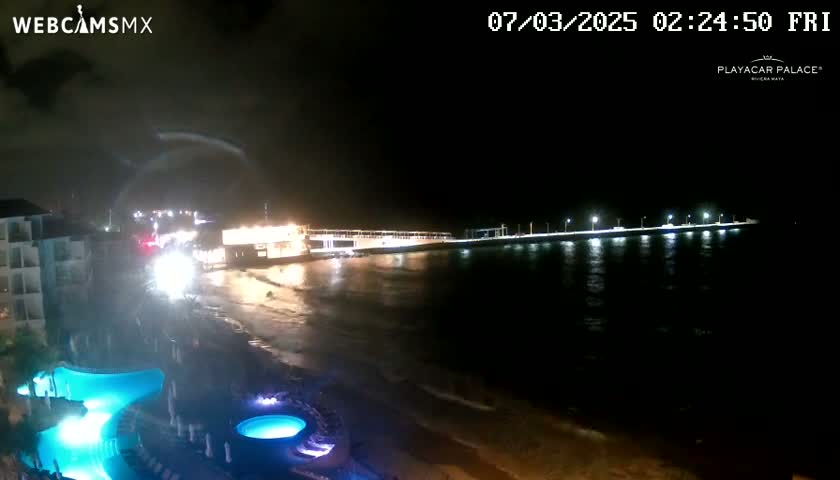 A nighttime beach scene shows a long, brightly lit pier extending into dark water, with a resort and illuminated pools in the foreground under partly cloudy skies.