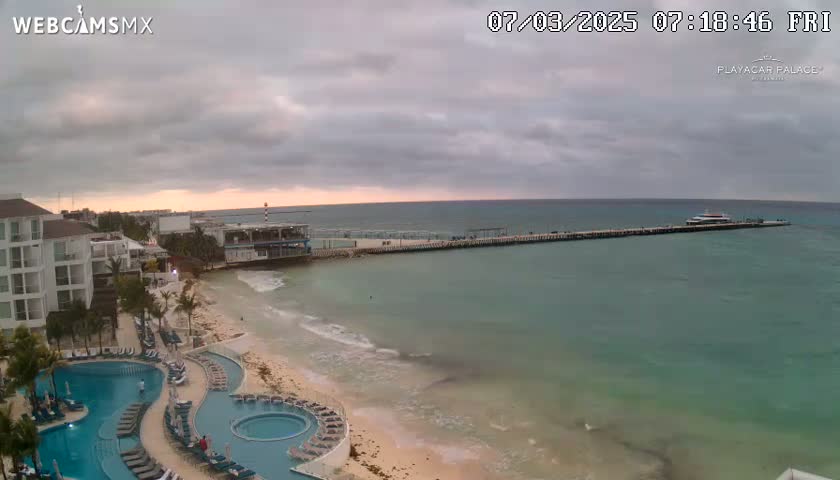 An overcast sky hangs over a beach scene with calm, turquoise water, a long pier, a resort with multiple pools, and a boat docked at the end of the pier.