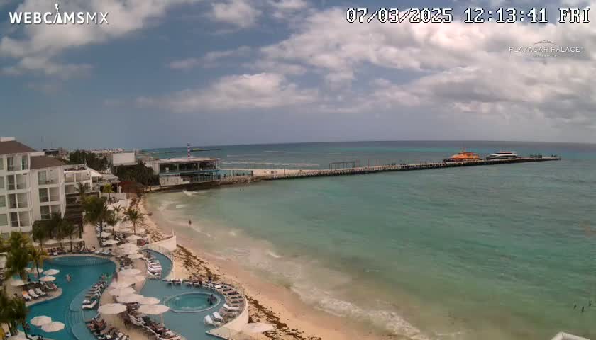 An aerial view of a resort beach on a partly cloudy day, showing a long pier with boats, turquoise water, and a sandy beach with multiple swimming pools.