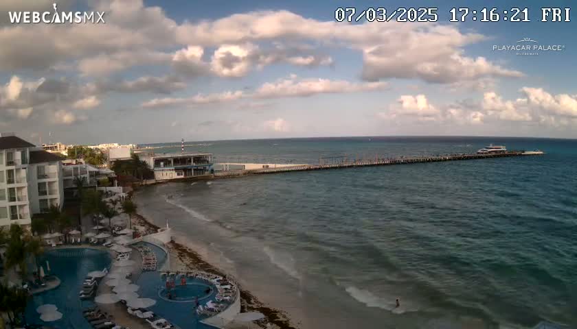 A partly cloudy afternoon scene shows a resort with a pool overlooking a beach, ocean, and pier with a boat.