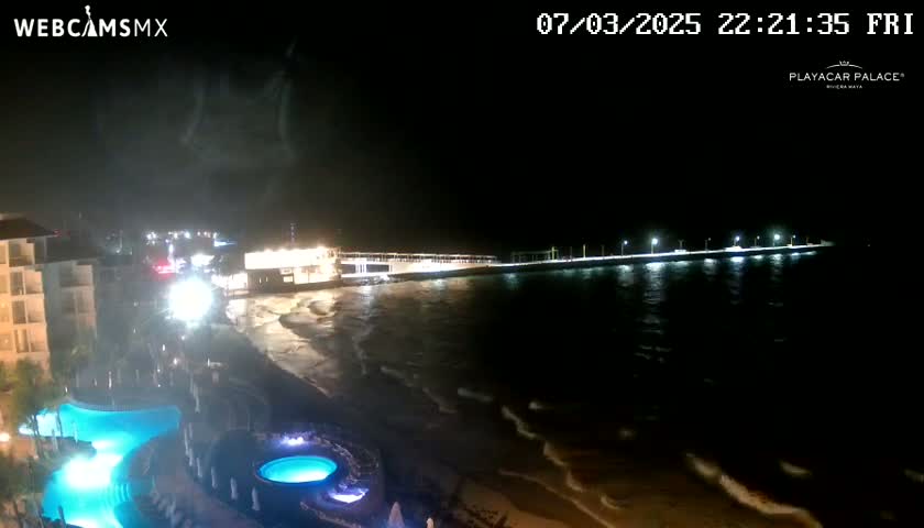 A nighttime view of a beach with a pier extending into the ocean, illuminated by lights, with gentle waves lapping the shore and a resort with illuminated pools in the foreground.