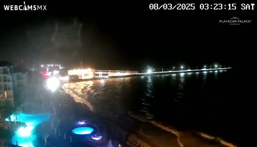 A nighttime view of a beach with a long pier lit up, gentle waves, and illuminated pools at a resort.