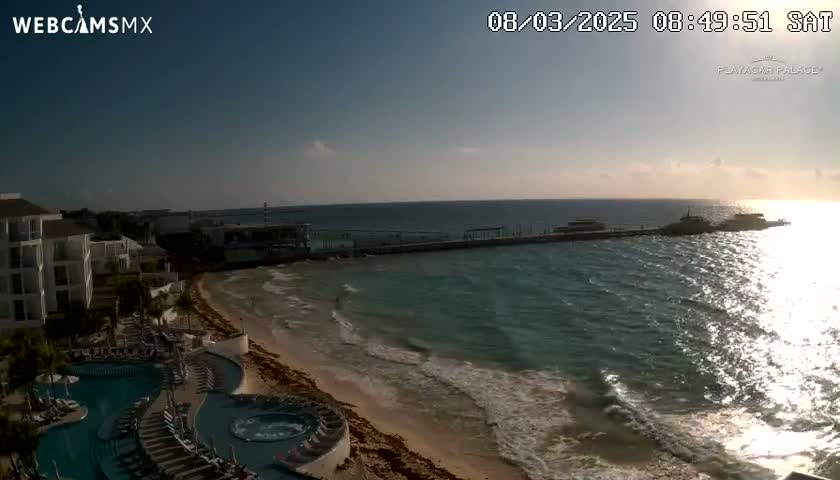 A beach with a resort, a pier, and calm ocean waves under a mostly clear sky during sunrise.