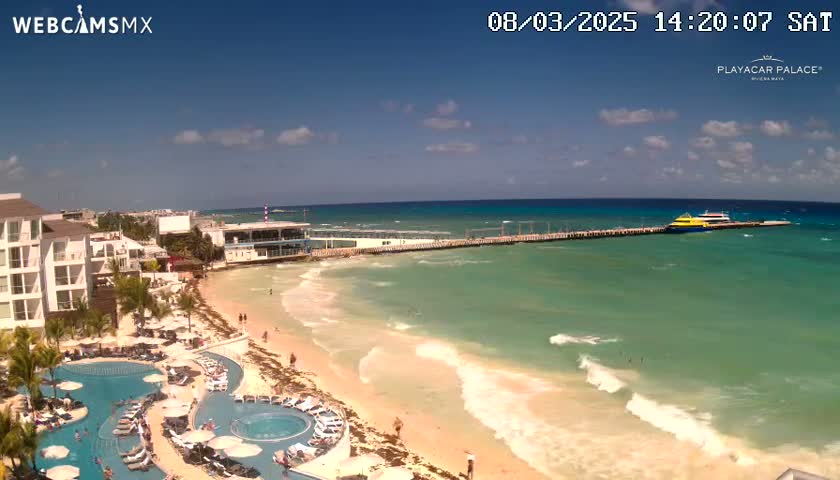 A sunny day shows a beach with turquoise water lapping gently on the shore, featuring a resort with pools and a pier with two boats docked.