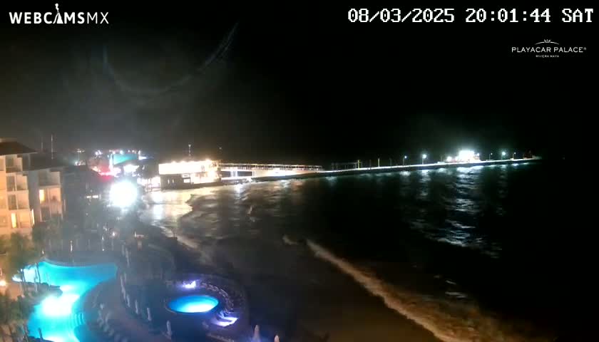 A brightly lit pier extends into a dark, wavy ocean at night, next to a resort with illuminated pools.