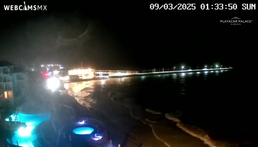 A nighttime view of a beach with a long, lit pier extending into the dark ocean, with gently rolling waves lapping the shore near a resort with illuminated pools.