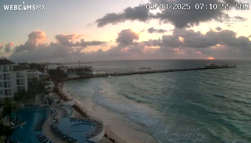 A sunrise with cloudy skies over a beach with a resort, pool, and pier extending into the ocean.