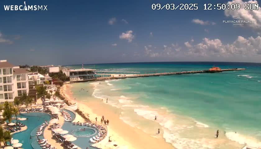 An aerial view shows a resort with multiple pools overlooking a long sandy beach and turquoise ocean under a partly cloudy sunny sky.