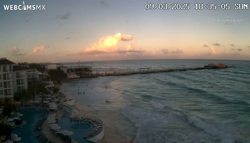 A resort overlooks a beach with calm waves at sunset, under a partly cloudy sky.