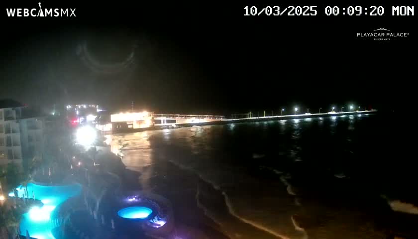 A nighttime view of a beach with a pier and lit buildings, showing gently moving water under a dark sky.
