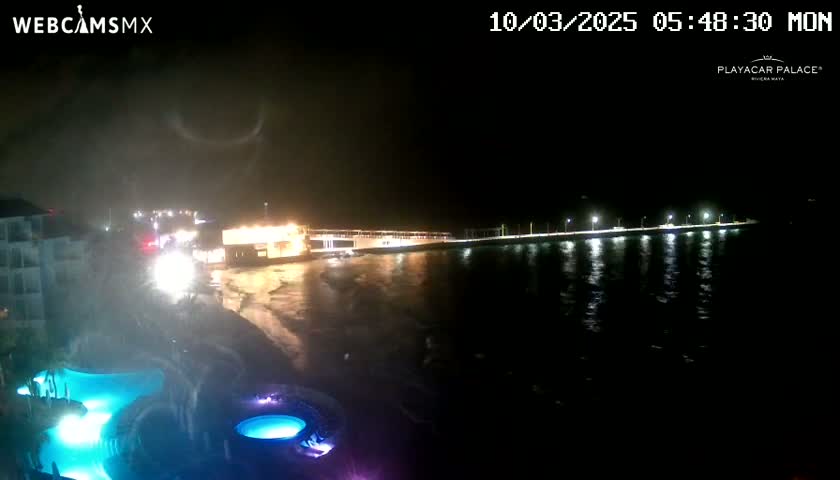 A nighttime view of a beach at a resort with illuminated pools and a pier extending into dark, wavy water under a cloudy sky.
