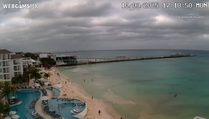 A resort area with pools and a beach overlooks a pier and calm, teal ocean under a cloudy sky.