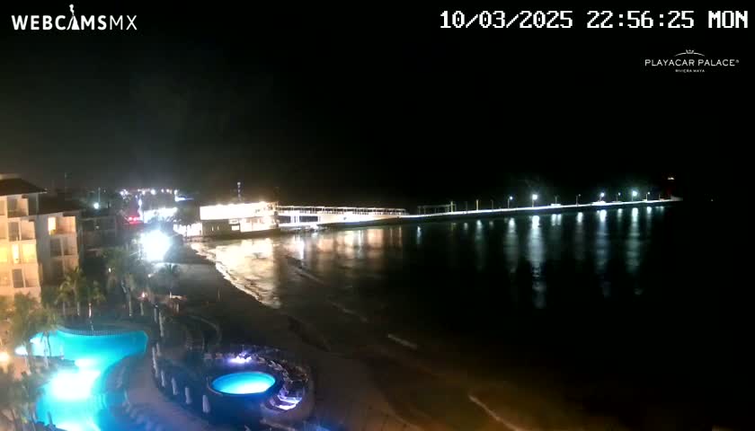 A nighttime view of a beach with a resort featuring illuminated pools, and a pier extending into the calm, dark water.
