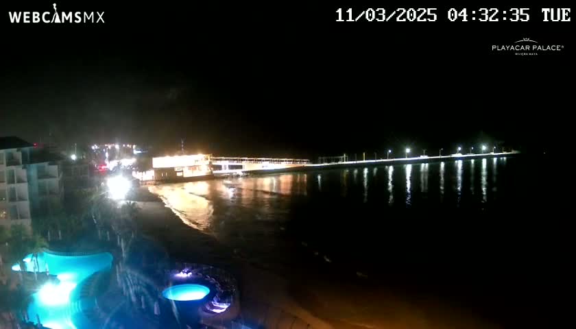 A nighttime coastal scene shows a brightly lit pier extending into dark water, with a building and swimming pools visible on the shore.