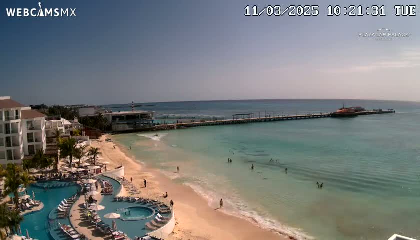 A beach resort with a pool area overlooks a calm, turquoise ocean with a pier and a boat visible in the distance under a clear sky.