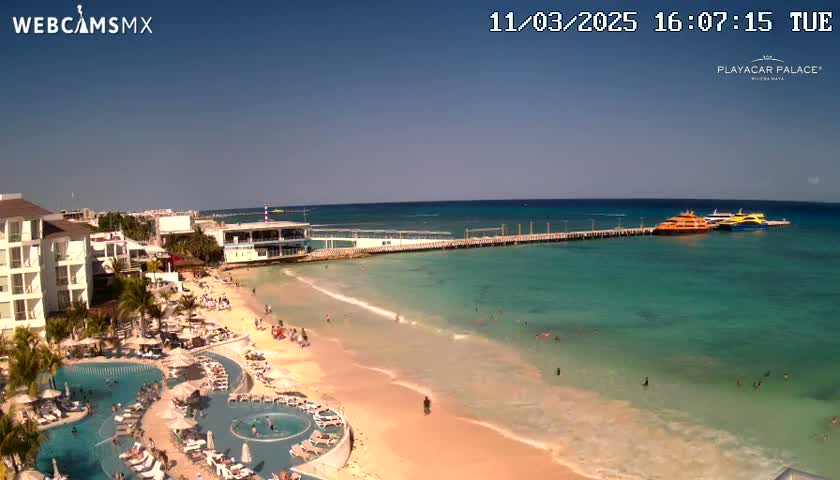 A beach resort with multiple pools overlooks a calm, clear turquoise ocean where several boats are docked at a pier under a sunny sky.