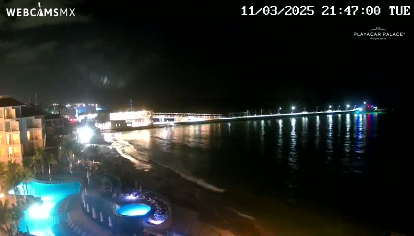 A nighttime view of a resort with illuminated pools overlooking a calm ocean and a brightly lit pier extending into the water.