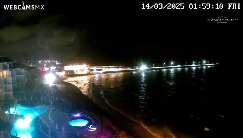 A nighttime view of a beach with a lit pier extending into the dark ocean, showing a resort with illuminated pools in the foreground under a partly cloudy sky.