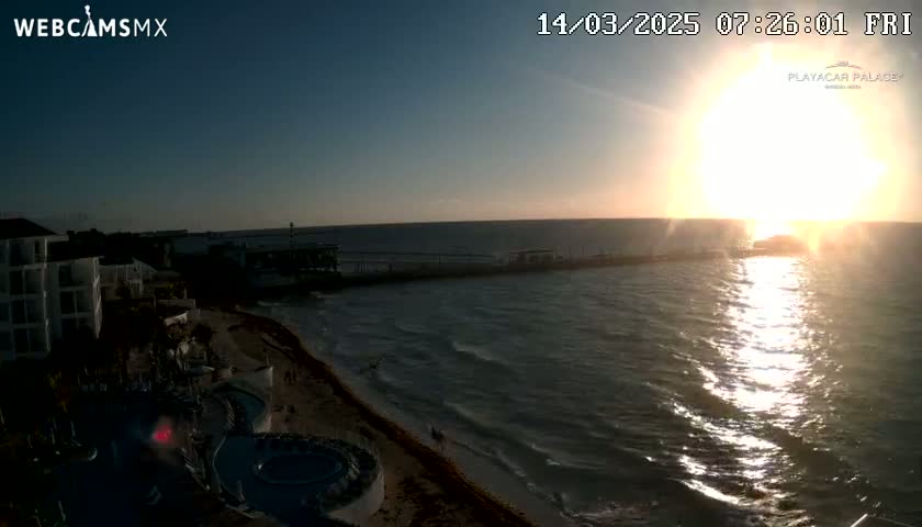 A coastal sunrise scene shows a beach with a resort, a pier extending into the ocean, with gentle waves and the sun shining brightly.