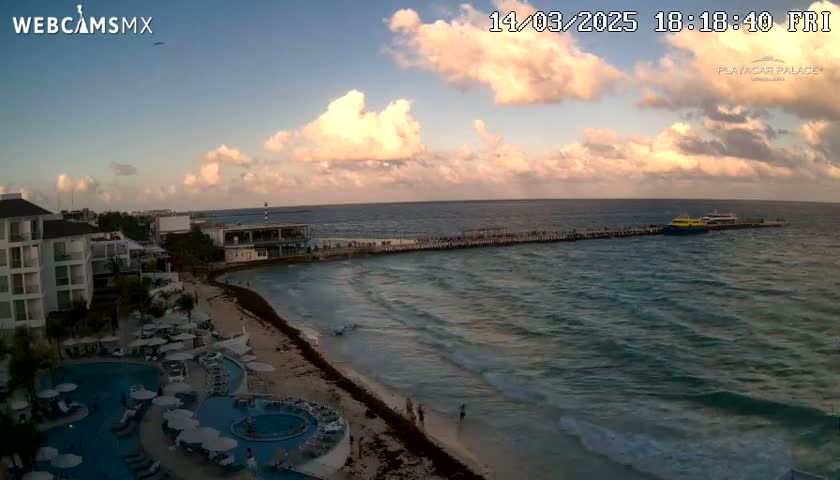 A partly cloudy sunset over a beach with a resort, a pier, and boats in the ocean.