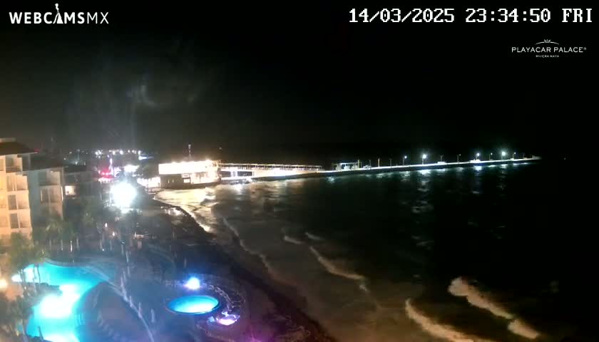 A nighttime coastal scene shows a resort with illuminated pools, a long pier extending into the ocean with gentle waves lapping the shore under a mostly clear sky.