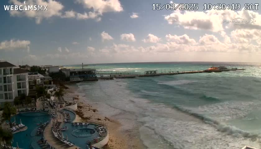 A partly cloudy day shows a resort with pools overlooking a beach and ocean with moderate waves, and a pier extending into the water with a boat docked at the end.