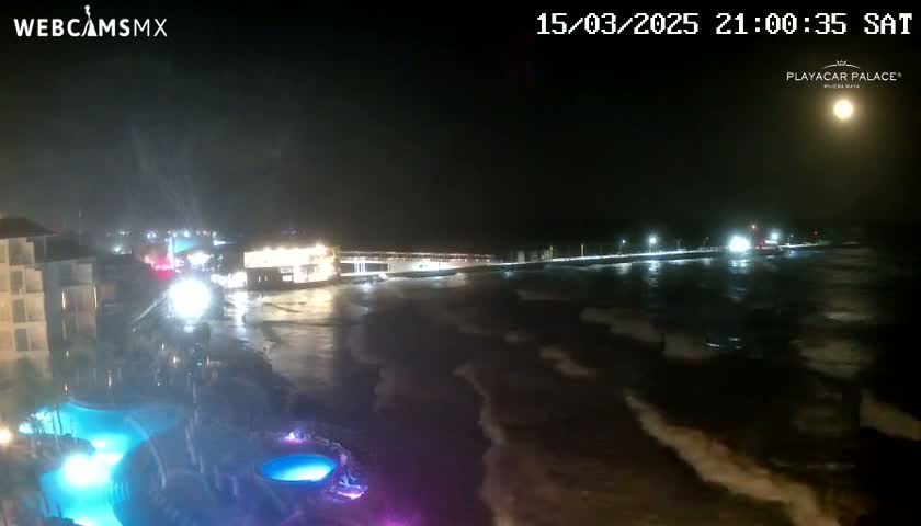 A nighttime view of a resort area with a pool illuminated in blue and purple, overlooking a dark ocean with waves under a bright moon.