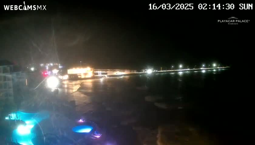 A nighttime coastal scene shows a body of water with waves lapping a shoreline where buildings and lights are visible, under conditions of heavy mist or spray.