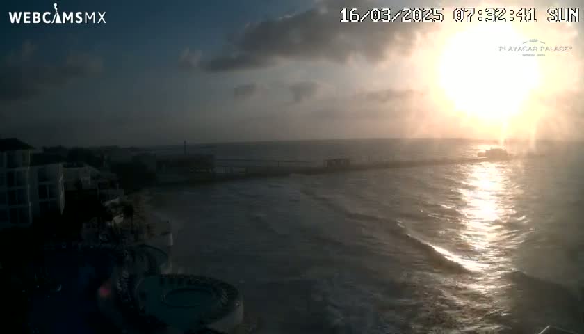 A sunrise over a wavy ocean with a pier and buildings in the background under a partly cloudy sky.