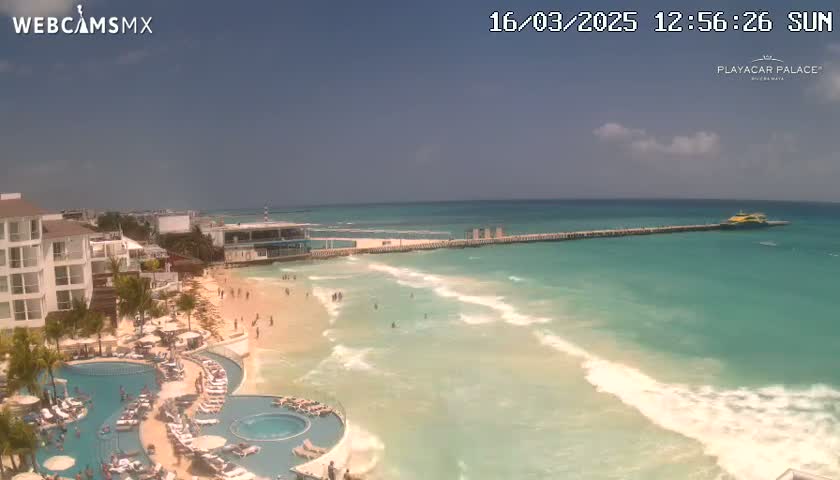 A sunny day at a beach resort shows a pier extending into a turquoise ocean with gentle waves lapping a sandy beach, where people are swimming and relaxing.