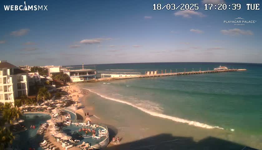A beach resort with a pool and pier extending into a calm, turquoise ocean under a mostly sunny sky.
