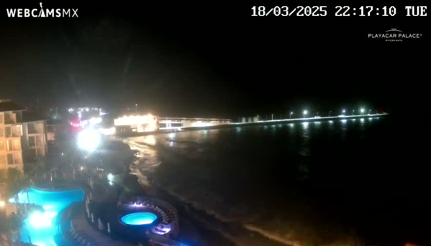 A nighttime view of a resort beachfront, showing a lit-up boardwalk stretching across the water, with calm waves lapping the shore near a pool area.