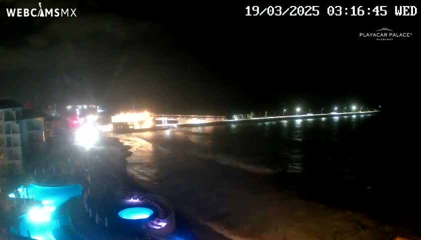 A nighttime view of a beach with a resort, illuminated pools, and a pier extending into dark water with gentle waves.