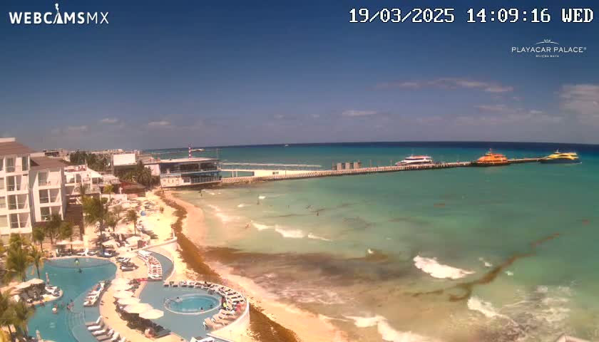 An aerial view shows a resort with pools and a sandy beach next to a clear turquoise ocean with waves, under a mostly sunny sky.