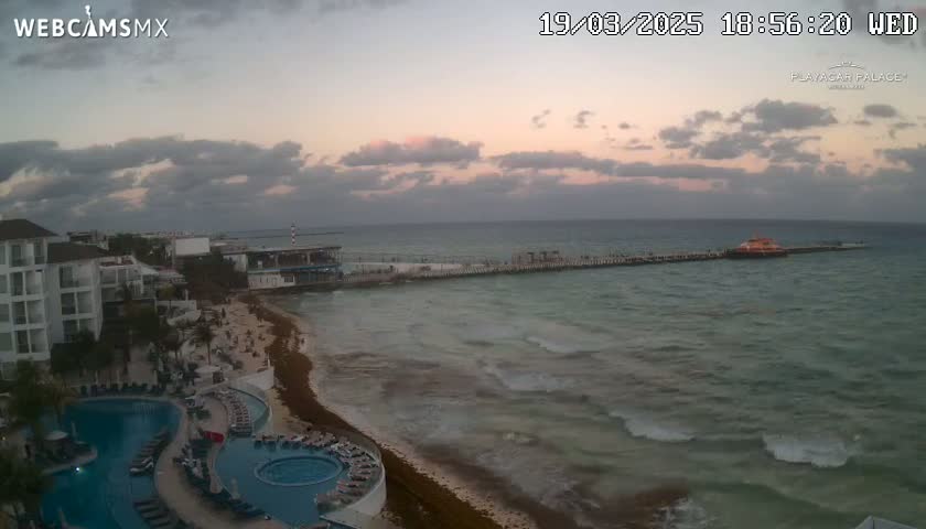 An ocean with moderate waves at sunset, a beach with seaweed, and a resort with swimming pools are visible from a high vantage point.