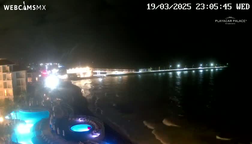 A nighttime view of a resort area shows a beach, ocean with small waves, a lighted pier, and resort buildings with illuminated pools.