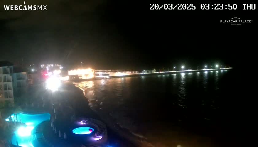 A nighttime view of a beach at a resort with illuminated pools and a pier extending into dark water.