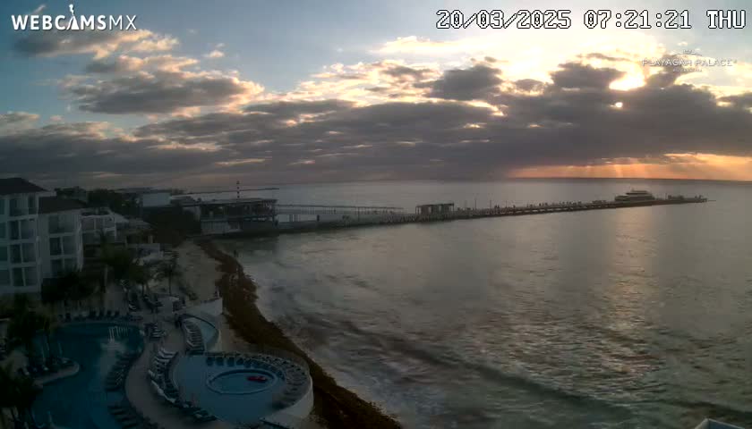 A partly cloudy sunrise illuminates a coastal scene featuring a resort with a pool, a long pier with a boat, and calm ocean waters.