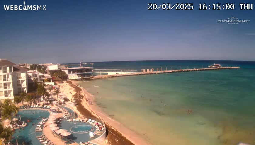 A resort with pools overlooks a beach and calm, turquoise ocean with a pier extending into the water on a clear, sunny day.