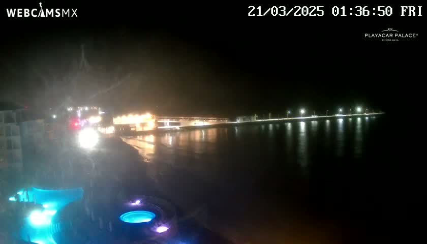 A nighttime view of a calm body of water reflecting lights from a shoreline building and streetlights, with illuminated pools in the foreground.