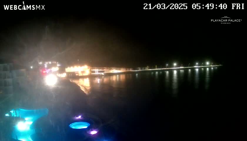 A nighttime view of a calm body of water reflecting lights from a shoreline with buildings and pools illuminated in various colors.