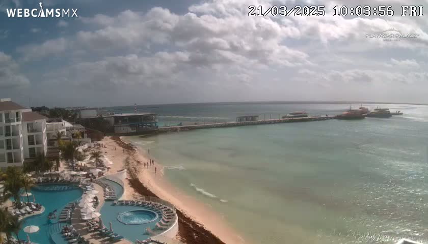 A partly cloudy day shows a resort with pools overlooking a sandy beach and calm ocean water with a pier and boats in the distance.