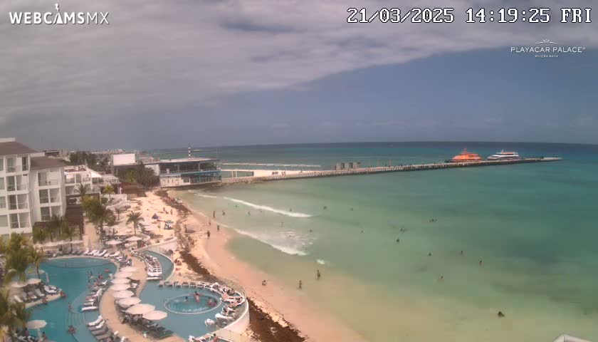 A beach resort with a pool area overlooks a partly cloudy sky, calm turquoise ocean, and a pier with boats docked at the end.