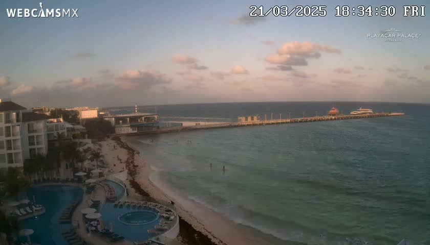 A beach resort with a long pier and two boats is seen at sunset under partly cloudy skies.