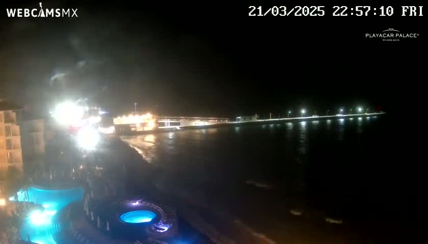 A nighttime view of a resort's illuminated pool area overlooking a calm, dark ocean with a brightly lit pier in the distance.
