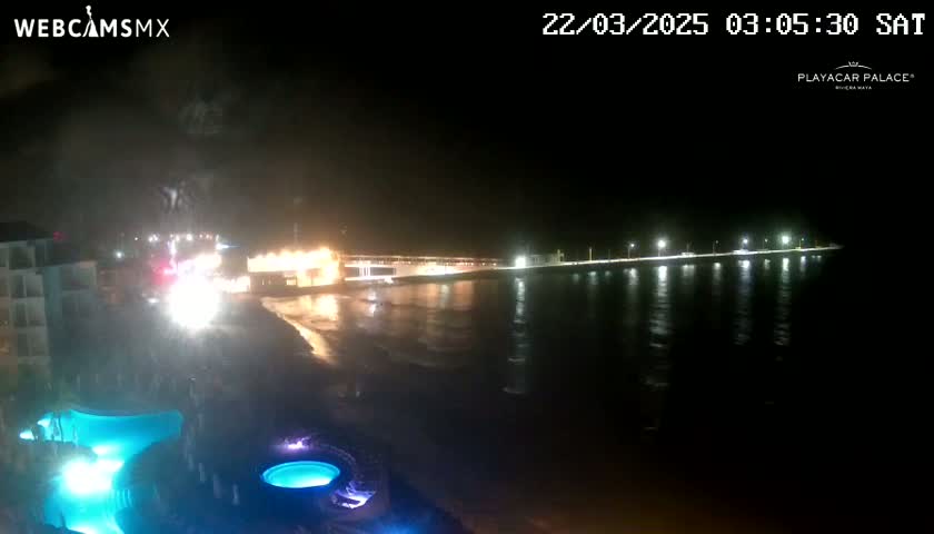 A nighttime view of a beach with illuminated buildings and pools in the foreground and a calm, dark body of water reflecting lights in the background.