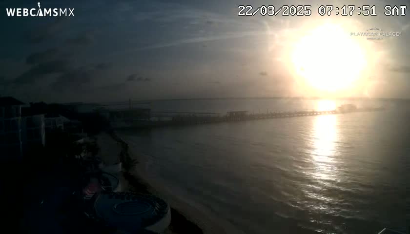 A hazy sunrise casts a golden glow over a calm ocean, illuminating a long pier extending from a shoreline with buildings and a swimming pool in the foreground.